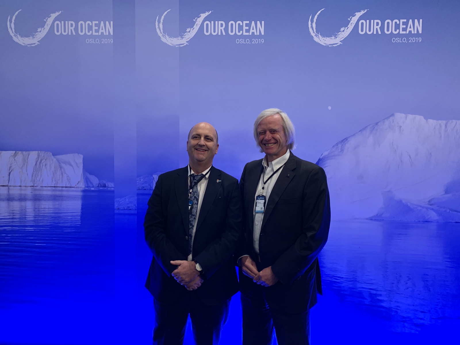 Two men in suits stand smiling in front of a blue backdrop featuring icebergs and the text "OUR OCEAN OSLO, 2019."