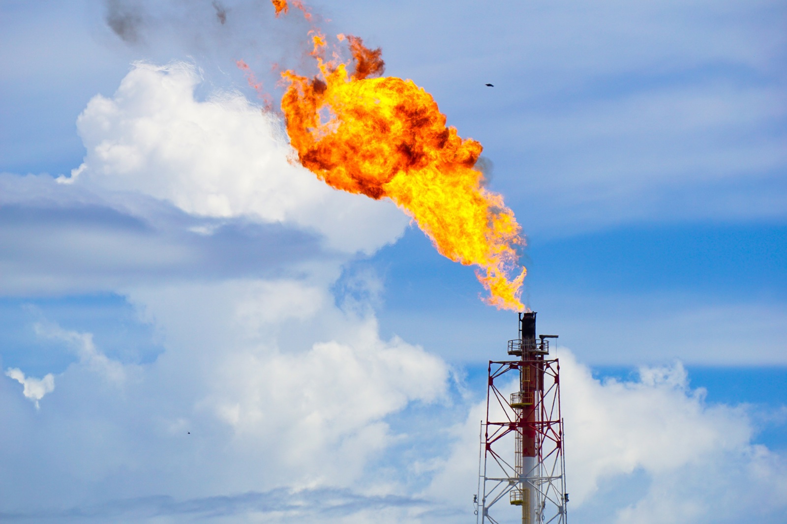 Industrial flare stack burning off excess gas with large flames against a backdrop of clouds and blue sky.
