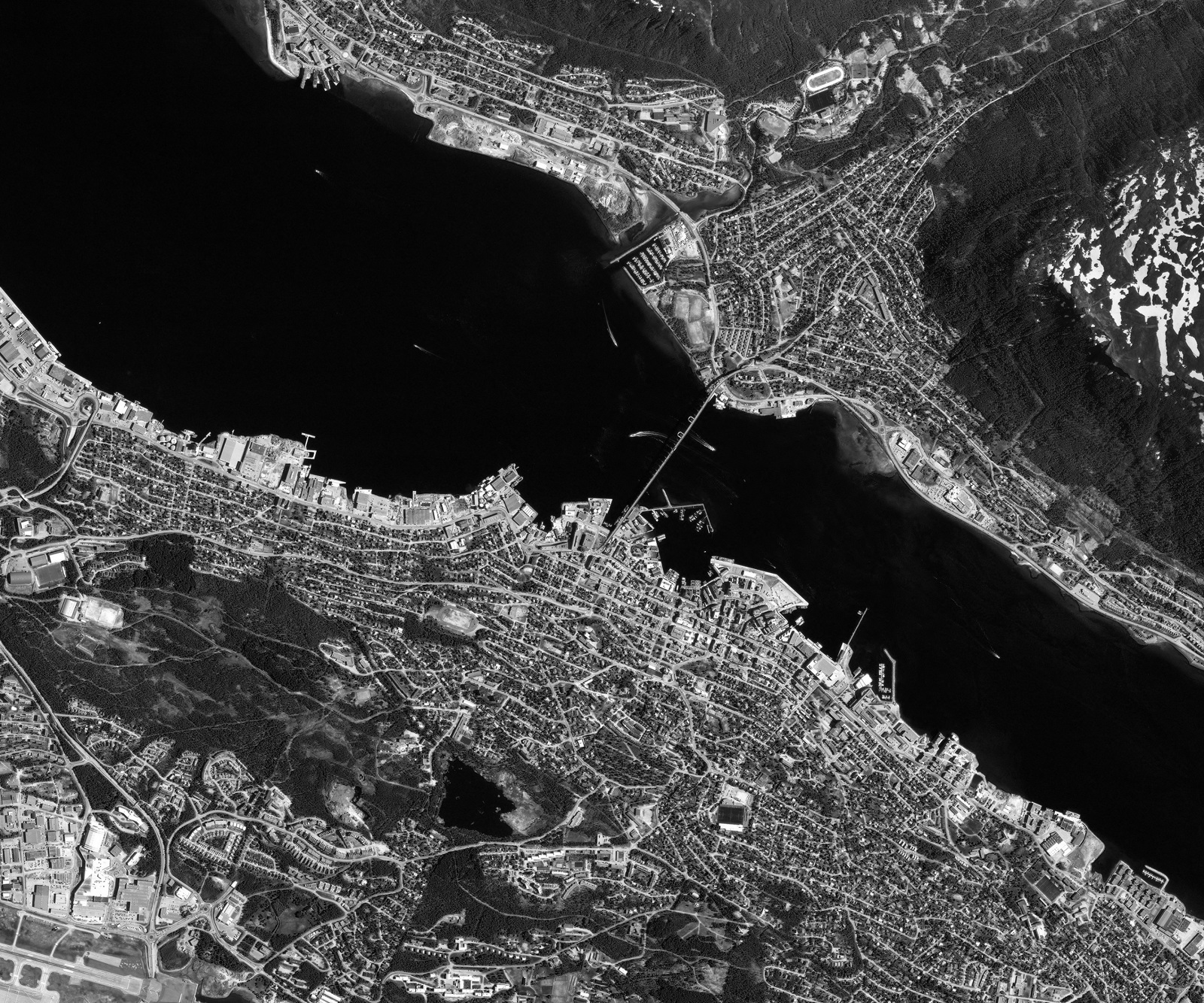 Black and white satellite image of a coastal city with dense urban areas, roads, and a body of water.