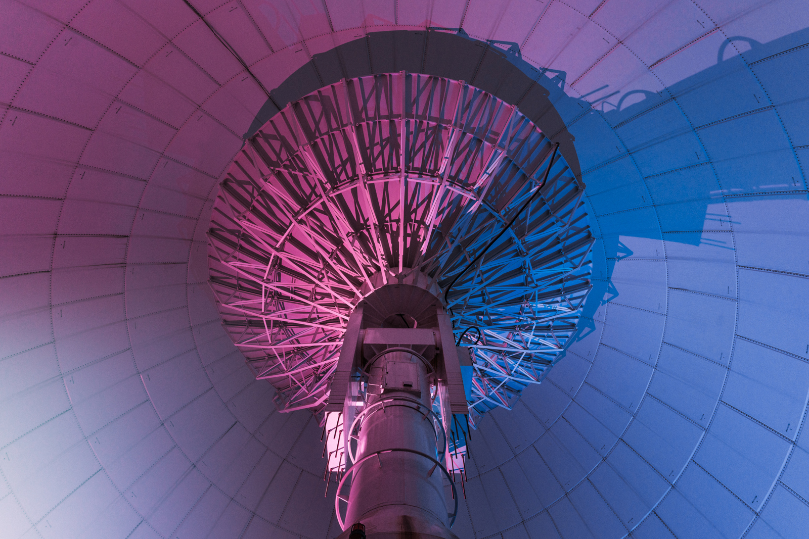 Interior view of a large satellite dish, illuminated with purple and blue lighting, showcasing its intricate structural details.