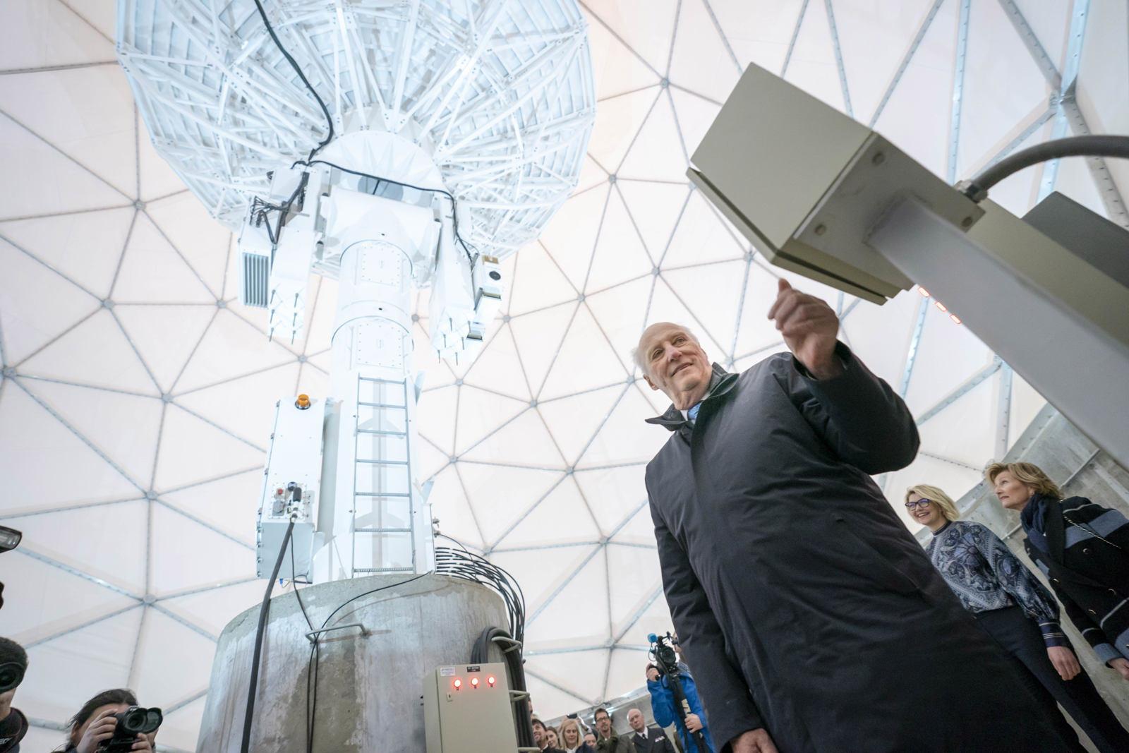 His majesty the King of Norway standing next to large satellite dish inside a dome structure, with several people and photographers in the background.